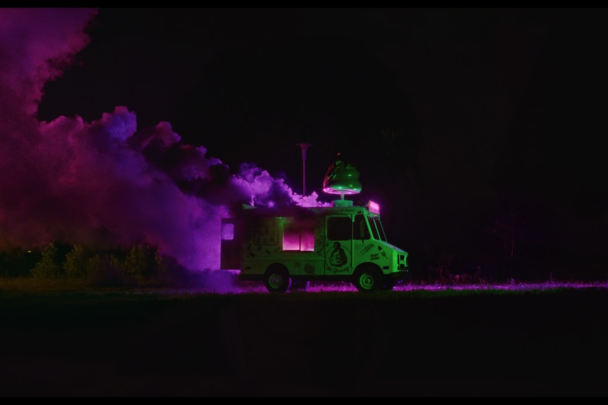 An ice cream van-style truck drives by at night with smoke billowing from it, tinted purple and pink by the light.