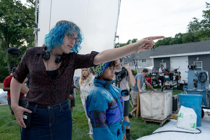 A person with shoulder-length blue hair points to something off-screen while sitting on a suburban US film set with a crew.